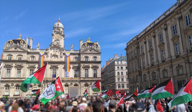 Foule portant des drapeaux palestiniens, devant l'hôtel de ville de Lyon, palais baroque portant la statue d'Henri IV à cheval et deux bannières LGBTQ+ "Lyon est fière !", et sous un grand ciel bleu.