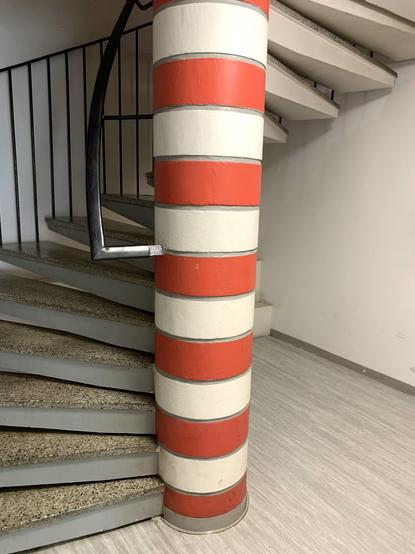 A spiral staircase with gray steps and a striped column painted in red and white. A black handrail curves around the column, leading upwards. The surrounding walls are white, and the floor has a light-colored surface.