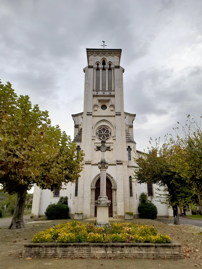 The outside of a light church in front of a dark sky. In front of the church is a cross on a pedestal between some yellow flowers. To the left and right are trees.