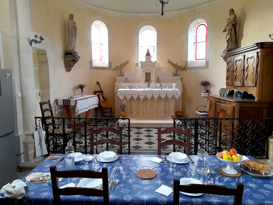 The inside of a former church with a table that is set in the front and a fridge to the left. Behind the table is an altar and some statues, as well as a guitar and a wooden cabinet.