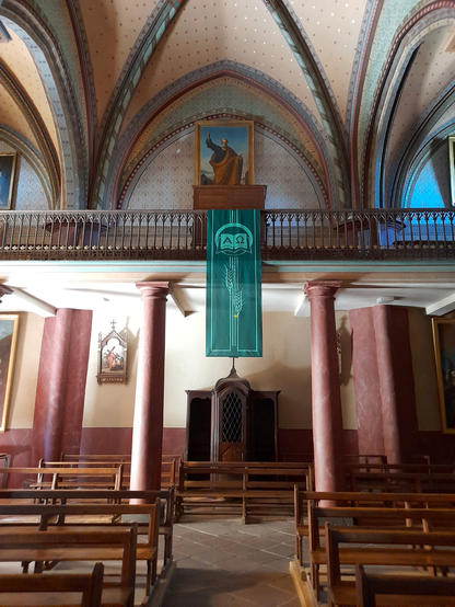 The inside of a church, looking towards the back, where a confessional is located in the middle behind the pews. The walls of the church are colourfully decorated in pastel colours and red and above the confessional a painting is hanging with a long blue banner in front of it.