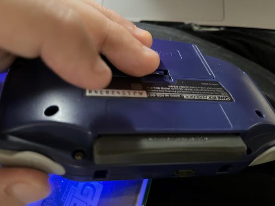 A hand is holding a blue handheld gaming device, showing the bottom side with buttons and ports visible. The device appears to be a Game Boy Advance. A GBA cart is inserted