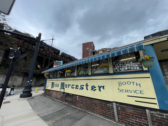 A diner, mostly yellow with a blue awning and trim. On the yellow facade is painted:

Booth Service (rounded letters)
Miss Worcester (fancy gothic letters)
Booth Service (rounded letters)

Seven pots of flowers hang from the awning.  We can see through the windows of the diner that they are covered on the inside with stickers and that there are white fluorescent tube lights on the ceiling inside.

To the left across the street is a dark steel train trestle, and atop that dark tanker railway cars. Both the trestle and cars have graffiti on them. The sky above is forebodingly cloudy and gray.