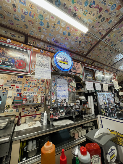 Interior of a diner, viewed from a counter seat. The beige wall and ceiling are busily covered by all different kinds of stickers. There is a shiny clock high on the wall with blue neon around it which has a picture of a yellow diner and say "Miss Worcester". The clock hands say 2:04. The whole view is chock-full of diner stuff: mik cooler, coffee maker and pots, cooler, spices, can of aerosol whip cream, bottle of chocolate sauce, below, coffee mugs. On the counter is a red ketchup bottle and other hot sauce bottles.

Two long fluorescent tubes are in a fixture in the ceiling.