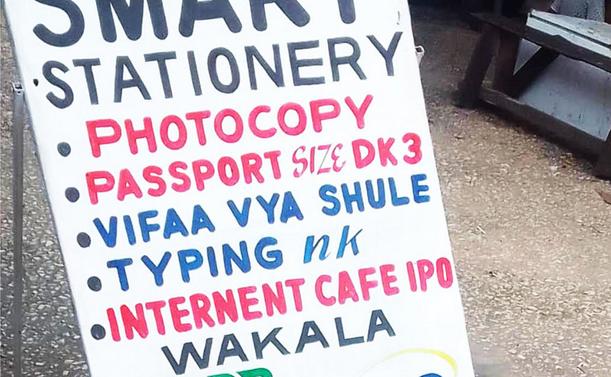 Hand-painted sign board for the Smart Stationery shop that contains a mixture of English and Kiswahili words to list the products and services available there:

Smart Stationery
Photocopy
Passport Size DK3
Vifaa Vya Shule
Typing nk
Internent Cafe IPO
Wakala