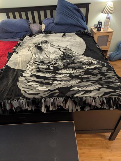 A fleece blanket with a tied fringe edge showing a very large and majestic wolf howling at a gigantic moon.