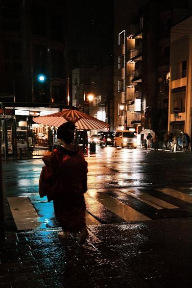 A geisha, or geiko, as they prefer in Gion, walks into a busy intersection facing away from the camera. She is carrying a parasol and wearing a red kimono. The streets are wet with rain and several buildings are brightly lit as cars pass by, headlights lighting her parasol.