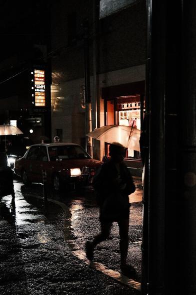 An individual hurries down a wet street wet with rain and glistening in the headlights of a cab. They are carrying a transparent umbrella, backlit by the warm light of a shop window across the street behind the cab.
