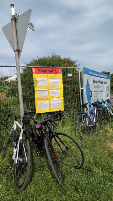 An einer Kontrollstation hängt ein großes Infoplakat an einem Bauzaun und mehrere Räder lehnen an einem Verkehrsschild.