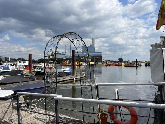 Blick auf eine kleine Marina mit mehreren angedockten Booten, umgeben von Wasser. Im Vordergrund ist eine Bogenkonstruktion mit dekorativen Elementen zu sehen, daneben ein Rettungsring. Der Himmel ist teilweise bewölkt, im Hintergrund sind Industriegebäude zu sehen.