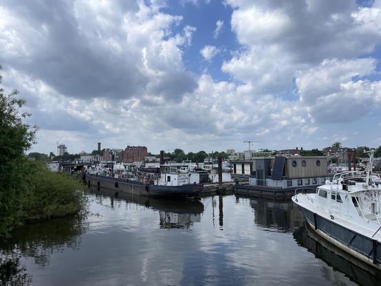 Blick auf eine kleine Marina mit mehreren Booten, die im ruhigen Wasser liegen. Im Hintergrund sind Gebäude und ein Kran unter einem teilweise bewölkten Himmel zu sehen. Auf der linken Bildseite ist üppiges Grün zu sehen.