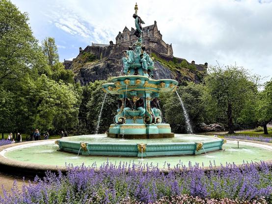 This image features the Ross Fountain, an elaborately decorated 19th-century cast iron fountain located in West Princes Street Gardens in Edinburgh, Scotland. The fountain is painted in vibrant turquoise and displays a variety of sculptures, including cherubs and mythical creatures. The fountain is set against the stunning backdrop of Edinburgh Castle which sits majestically on a hill above the gardens. The surrounding area is lush with greenery and blooming purple lavender, adding a colorful contrast to the scene. This picturesque setting is popular among both locals and tourists.