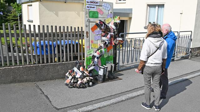 Zwei Personen stehen auf einer Straße und betrachten eine alte Tür, die an einem Holzzaun befestigt ist. Vor der Tür ist schräg ein Kreuz aus Schuhen festgemacht. Auf der Tür und auf dem Kreuz kleben Zettel mit Botschaften zum Nachdenken.