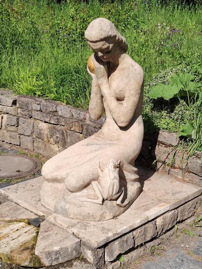 Eine Prinzessin aus Stein. In der Hand hält sie eine goldene Kugel. An ihrem Bein sitzt ein Frosch und heischt um Aufmerksamkeit.