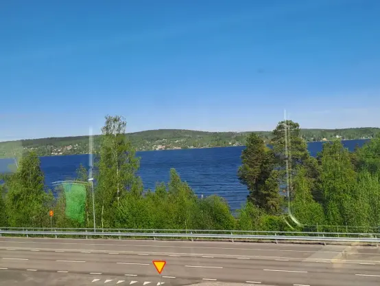Photograph (colour): The Baltic Sea, seen from a train window. In the front a street and trees, in the back a wood covered hill