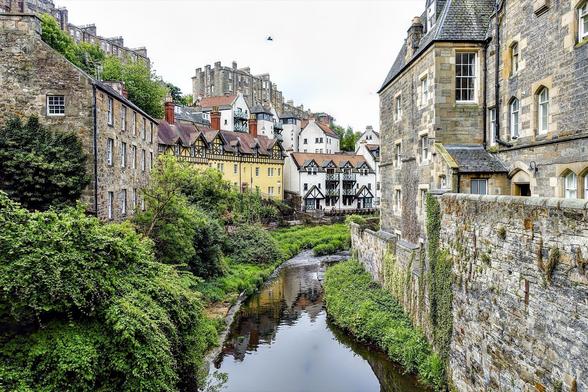 This image showcases a picturesque scene that appears to be from a historic village or town. Key features include:

- A river flowing gently through the middle, lined with lush greenery on its banks.
- A variety of buildings, each with unique architectural styles that suggest a mix of different historical periods. On the left is a row of multistory stone buildings, possibly used as residences or shops.
- On the right, a part of the scene includes buildings with a very distinctive Tudor style, characterized by exposed wooden frameworks and white plaster.
- The background features additional buildings on elevated terrain, possibly indicating that this area is in a hilly or mountainous region. The architecture here also varies, adding to the location's charm.
- The entire scene is overcast, suggesting a cool or mildly damp climate, which is common in many European historical towns.
- There's also a bird flying in the sky, which adds a dynamic element to the serene setting.

This image likely captures the essence of a European medieval town, possibly in the UK, given the architectural styles and natural surroundings and is named Dean Village in Edinburgh.
