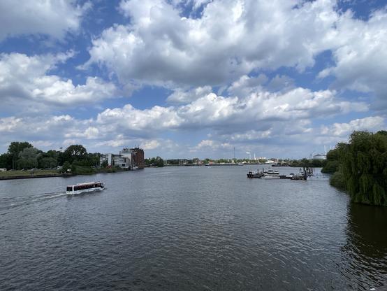 Eine Flussszene mit einem Amphibienfahrzeug auf dem Wasser, üppigem Grün und Gebäuden entlang der Uferlinie. Der Himmel ist teilweise bewölkt, was eine ruhige Atmosphäre schafft.