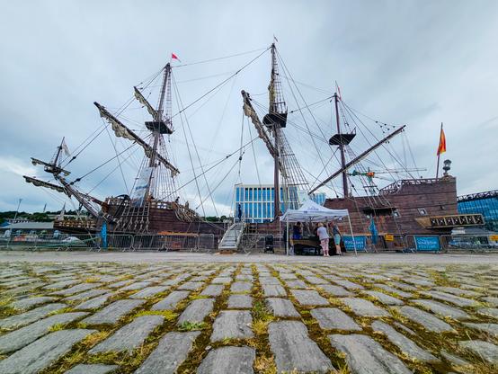 A wide-angle photo taken at ground level in Cork, Ireland, showing a large replica of a historic wooden sailing ship docked at a harbour festival. The ship features three tall masts with rigging and furled sails, a Spanish flag at the stern, and several people gathered under a white tent near the gangway. The foreground is cobbled stone pavement with grass growing between the stones. Signs read "HARBOUR FESTIVAL."