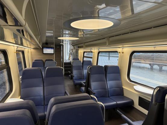 Interior of the lower deck of a double decker train. Big windows left and right and in the middle blue seats visible and dark colored floor. In the ceiling circular lights are visible. No seat is occupied.