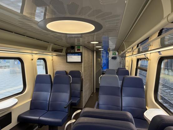 Interior of the lower deck of a double decker train. Big windows left and right and in the middle blue seats visible and dark colored floor. In the ceiling circular lights are visible. No seat is occupied.