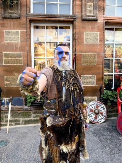 The image shows a man dressed in a dramatic, historical or fantasy-inspired costume portraying a warrior. He has his face painted with blue designs, possibly to mimic ancient or tribal war paint. He is wearing attire that includes animal furs and a leather tunic, accessorized with ropes, beads, and a sword attached to his belt. He is also holding what seems to be a mug or a similar object and striking a playful pose extending his fist toward the camera. In the background, there are buildings with stone and brick facades, suggesting the photo might have been taken in an urban area rich in historical architecture, possibly during a festival or a reenactment event.
