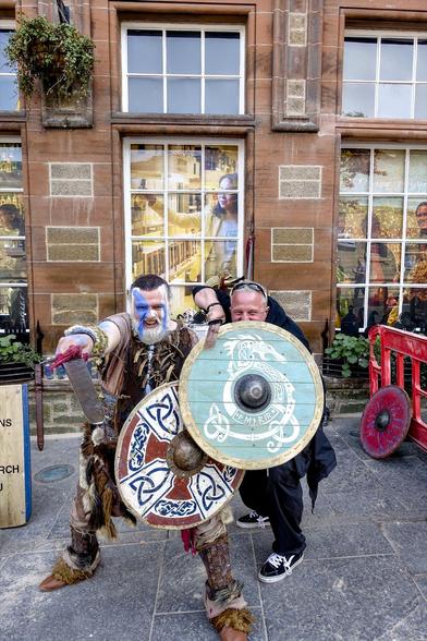 This image shows two people dressed in Viking-themed costumes. The person on the left is wearing elaborate attire with detailed face paint resembling a Viking warrior and is holding a sword and a shield adorned with Nordic symbols. The person on the right, partially obscured, also appears to be supporting the left person in a playful or demonstrative pose. The setting looks to be a public street in front of a stone building with large reflective windows that display bystanders watching the scene. The encounter captured in the photo gives it a lively and festive feel, likely part of a historical or cultural reenactment event or a festival celebrating Viking history.