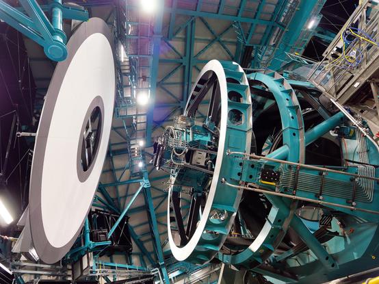 Rubin Observatory's huge 8.4-meter telescope points at the in-dome calibration screen. On the right, and tilted left nearly to horizontal, the telescope is a huge, teal steel structure with a top and middle ring supported by thick beams connected to the telescope base. On the left, the calibration screen is a large white donut shape slightly wider than the top end of the telescope. Credit: RubinObs/NOIRLab/SLAC/DOE/NSF/AURA/W. O'Mullane