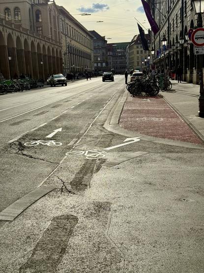 Stadtstraße mit Fahrradweg-Markierungen, abzweigendem Radstreifen und parkenden Fahrrädern. Im Hintergrund historische Gebäude und wenige Autos bei tiefstehender Sonne.