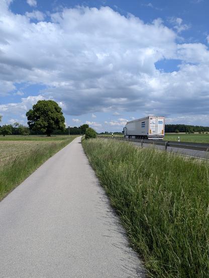 Une piste cyclable goudronnée longe une route départementale (Landstrasse) sur laquelle se situe un camion. La piste cyclable est sur le coté gauche de la route et elle est en est séparée par une bande d'herbes hautes.