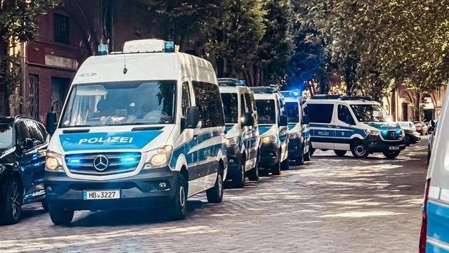 Eine detaillierte Aufnahme zeigt eine Kolonne von mehreren modernen weißen Polizeibussen mit blauen Streifen auf einer leicht sonnigen, gepflasterten Straße. Mindestens sechs Busse sind sichtbar, in einer Linie aufgereiht. Jeder Bus trägt deutlich lesbar die Aufschrift "POLIZEI" an den Seiten und vorne. Die Fahrzeuge, erkennbar als Mercedes-Benz Sprinter, sind mit Blaulicht ausgestattet, das bei einigen Fahrzeugen aktiv leuchtet und für eine dynamische Wirkung sorgt. Der vorderste Bus, der im Fokus steht, zeigt das Kennzeichen "HB 3227", was auf Bremen hinweist. Die Busse stehen dicht beieinander, was den Eindruck eines geschlossenen Verbandes oder Konvois vermittelt. Die Straße wird von Bäumen mit vollem Laub und teils historischen Gebäuden gesäumt, deren Architektur im Hintergrund angedeutet ist. Weitere zivile Fahrzeuge sind am Straßenrand weiter hinten geparkt, was auf eine belebte Umgebung schließen lässt. Die Komposition des Bildes lenkt den Blick entlang der Fahrzeugreihe in die Tiefe der Straße und betont die Länge des Konvois. Die Beleuchtung mit langen Schatten suggeriert spätes Tageslicht. Das Bild vermittelt die Präsenz und Mobilisierung von Polizeikräften, möglicherweise im Rahmen eines Großeinsatzes, einer Patrouille oder der Verlegung von Einsatzkräften in einem urbanen Gebiet. Die Szene strahlt eine Atmosphäre der Ordnung und potenziellen Einsatzbereitschaft aus.