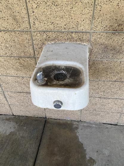 A white porcelain drinking fountain on the tan brick wall of the clubhouse. The basin is black with dirt. This is an improvement —a week before this the basin was clogged & overflowing.