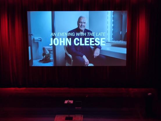 An empty stage with a still picture. Caption: an evening with the late John Cleese