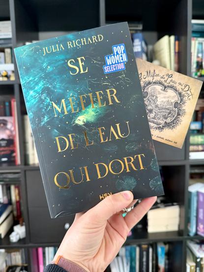 Livre pris en photo devant une bibliothèque avec un frontispice dessiné glissé dans ses pages. 
La couverture de Se méfier de l’eau qui dort montre un personnage aux traits féminins, allongé dans une rivière un peu agitée. Des particules dorées parsèment la page et accompagnent la dorure brillante du titre et du nom de l’autrice.