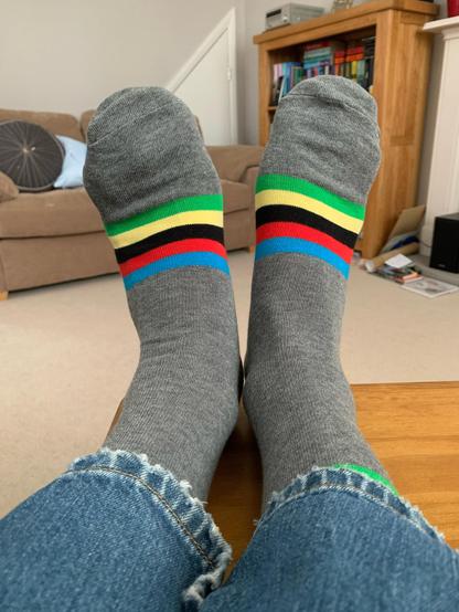 Two feet in socks, propped up on a small table. The view is from the wearer's perspective. The socks are grey with rainbow coloured bands around the instep, starting with blue, then red, black, yellow and green.