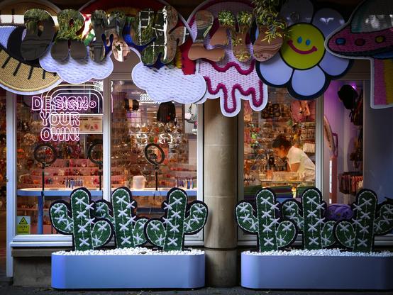 A very colourful shop in Neal Street, in London's Covent Garden area. A lady shop assistant can be seen inside the shop at the right, leaning on the counter and checking her phone while waiting for customers. Hundreds of colourful gifts fill the shelves and the walls throughout the shop. Above the windows outside are shapes made of fabric including a large eye, clouds, a daisy, and a UFO. Below the windows are six fabric cactuses standing in two light blue planters full of white-painted gravel.