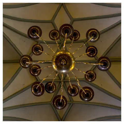 quadratisches bild mit weißem rand:
ein von unten fotografierter kronleuchter mit vielen rundungen, die auf diversen armen abgehen. dahinter ein muster der kirchendeckenkonstruktion