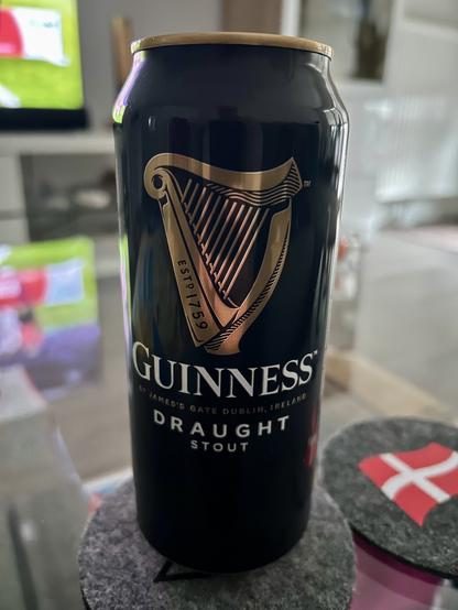 Auf dem Bild ist eine schwarze Dose Guinness Draught Stout zu sehen. Die Dose steht auf einem runden Filzuntersetzer, der auf einem Glastisch platziert ist. Auf der Vorderseite der Dose ist das bekannte goldene Guinness-Harfen-Logo prominent abgebildet, darunter steht in großen weißen Buchstaben „GUINNESS“ und darunter „DRAUGHT STOUT“. Im Hintergrund erkennt man unscharf einen Fernseher, auf dem offenbar ein Sportereignis läuft, sowie weitere Einrichtungsgegenstände eines Wohnzimmers. Rechts neben der Dose liegt ein weiterer Filzuntersetzer mit einer dänischen Flagge. Die Szene wirkt gemütlich und deutet auf einen entspannten Abend, vermutlich beim Fernsehen, hin.