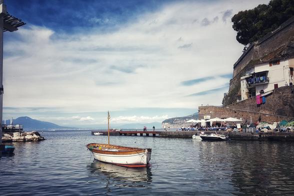 A small wooden boat floats on calm water in the Marina Grande in Sorrento, Italy, with a bustling restaurant and cliffside buildings in the background under a partly cloudy sky.