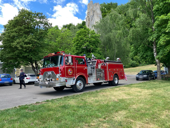 A vintage red firetruck from 1982, displayed outdoors for the 150th-anniversary celebration of the volunteer fire department in Wellheim. In the background, the prominent rocky cliff “Dohlenfelsen” rises amidst lush greenery, highlighting a popular climbing area.