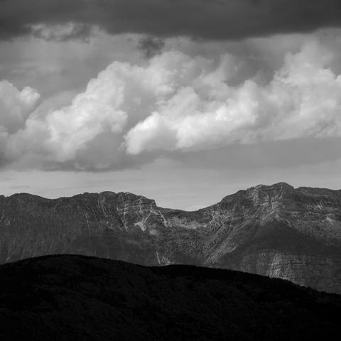 Photo noir et blanc d'un paysage.
En premier plan, des collines boisées dans l'ombre.
Puis de hautes falaises avec quelques névés au soleil.
Enfin de ciel chargé de nuages bourgeonnants.