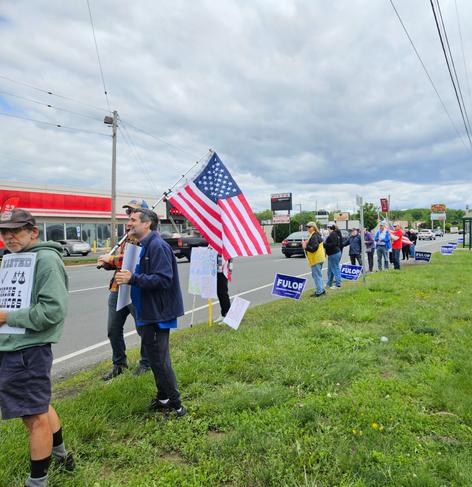 A group of protesters in Springfield, NJ holding signs, including an American flag, stands along a roadside with businesses in the background under a cloudy sky. Image by ‪Jim Cardillo‬
 ‪@jimcardillo.bsky.social‬