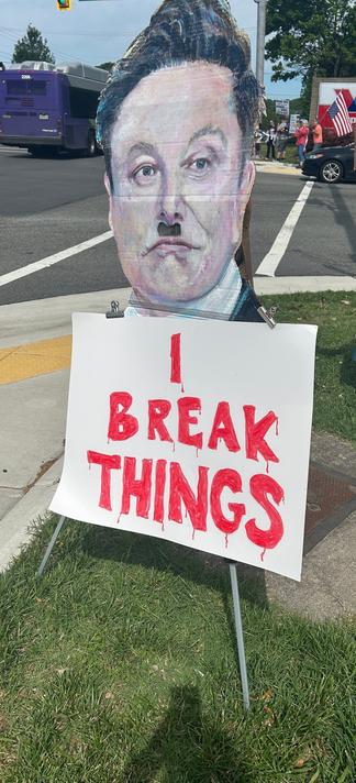 A sign reading "I BREAK THINGS" in bold red letters stands on grass beside a busy road, with cars and a bus passing by. Image by 
‪Ericka Esquire‬ ‪@erickaesquire.bsky.social‬