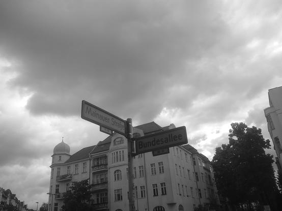 A black and white image depicting a street sign at the intersection of Mainauer Straße and Bundesallee. The background features a cloudy sky and nearby buildings with architectural details.