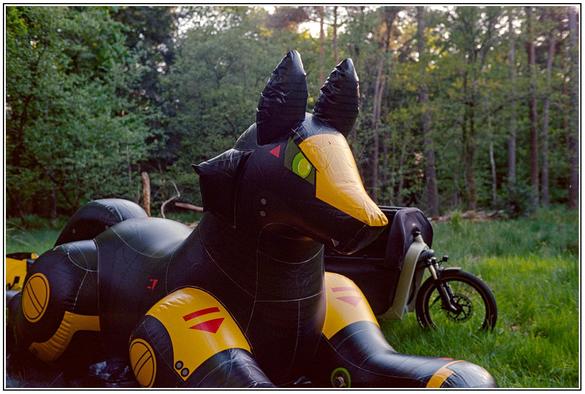 A big inflatable mecha looking fox lying in the forest behind it a cargo bike is parked, the colors moody and dark