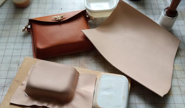 A work bench with a rectangular leather bag on it. The bag is British tan with a bee closure. There is also a modest sized piece of vegetable tanned leather on it. The top center shows part of a plastic food container. The bottom shows a rectangular piece of leather, shaped like a box and stapled to a pine board. A plaster mould is to the right to it.