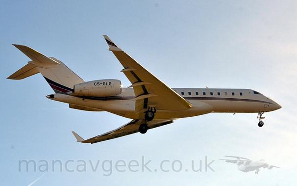 Low side view of a white, twin engined Bizjet flying from left to right at a very low altitude.
The plane has it's undercarriage lowered, and flaps extended from the rear of the wings, suggesting it is about to land.
The plane is mostly white, with a dark blue, red, and grey stripe running along the body from the nose, getting slightly wider and sweeping up in to the lower reaches of the tail.
A similar stripe is on the outer face of the up-turned wing-tips.
Bright but hazy sky with hints of blue fill the rest  of the frame.