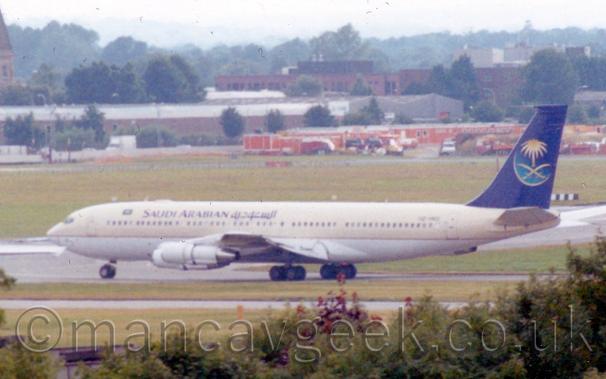Slightly grainy and blurry side view of a creamy-coloured, 4 engined jet airliner taxiing from right to left,
The plane has creamy upper surfaces, and a white belly.
There are dark blue "Saudi Arabian" titles on the upper forward fuselage.
The tail is the same dark blue, with a pale blue crescent moon, crossed golden sabres, and golden palm tree.
Trees and bushes line the bottom of the image, with grass leading up to buildings and trees in the distance.