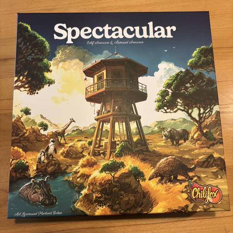 Das Bild zeigt die Oberseite eines Brettspielkartons mit dem Titel "Spectacular". Das Bild zeigt eine malerische Landschaft mit einem Wachturm, einer Vielzahl von Tieren, darunter eine Giraffe, ein Nashorn, ein Pinguin, ein Schuppentier und ein Nilpferd.