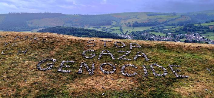 On a sloping grassy rampart, overlooking valley far below and a small town to the right, in big letters made of rocks, the message "Stop Gaza Genocide"
