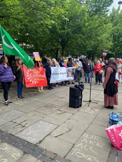 Protestors are assembled holding various banners including "UNITE COMMUNITIES". They are surrounding a war memorial, in front of which is a sound system and standing mic. A Muslim lady in hijab is delivering a speech. The paved area in front of her is covered in chalk murals with messages of love and hope, including "WE ARE TOGETHER".